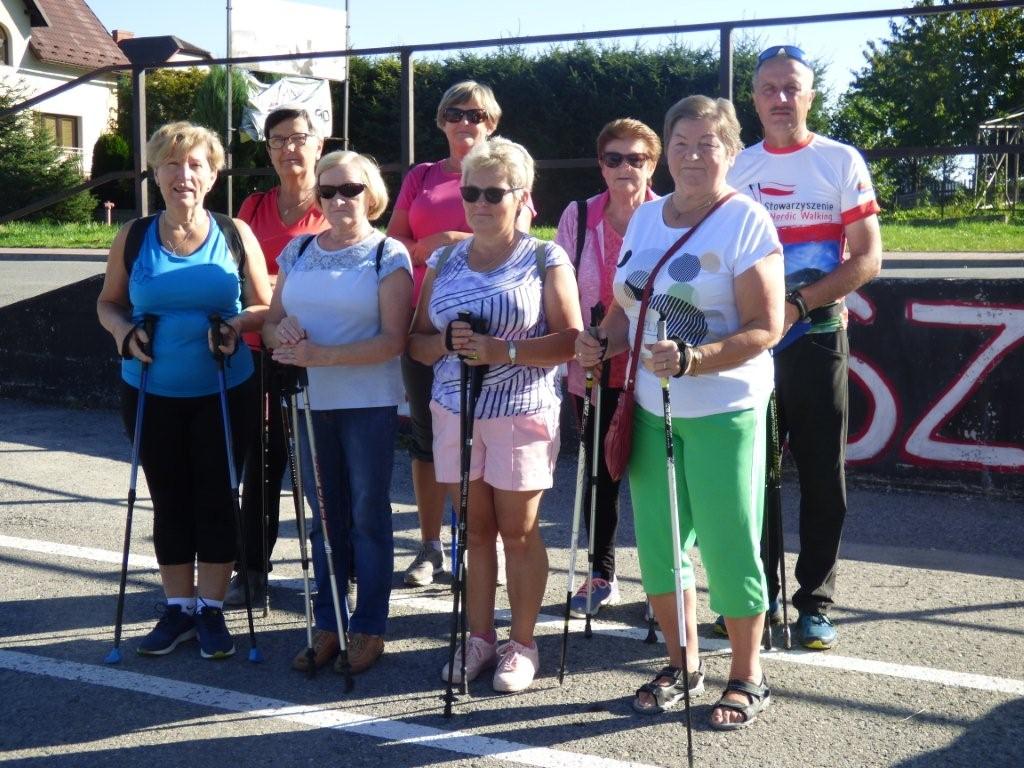 Seniorki w strojach sportowych z kijkami nordic walking stoją razem z instruktorem i pozują do grupowego zdjęcia.