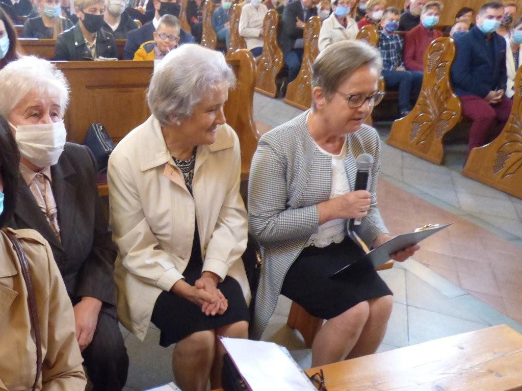 Trzy seniorki siedzą na ławce, jedna z nich trzyma mikrofon i czyta wiersz.