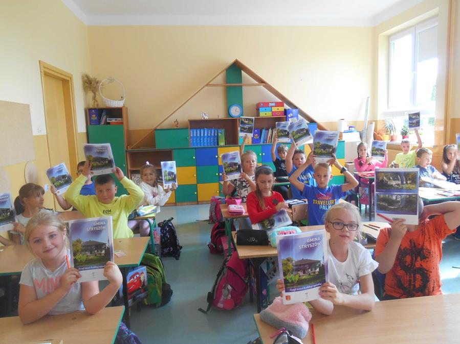 Dzieci z Łękawicy siedzą w ławkach w klasie i pozują do zdjęcia trzymając zeszyty ćwiczeń w rękach.
