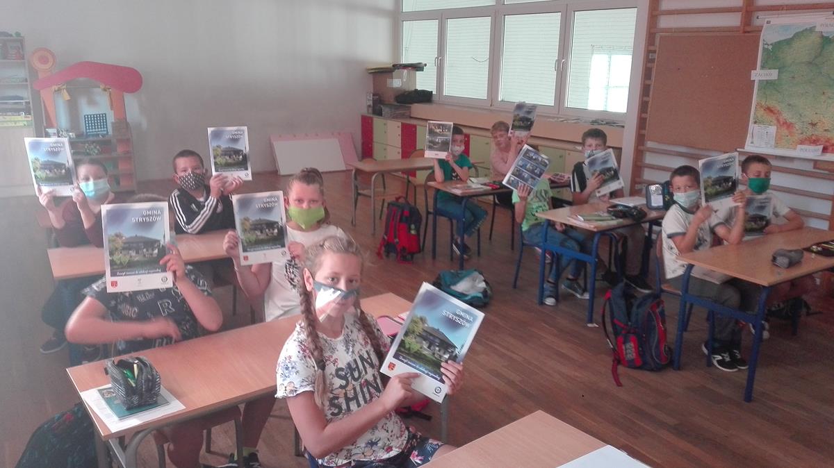 Dzieci z Łękawicy siedzą w ławkach w klasie i pozują do zdjęcia trzymając zeszyty ćwiczeń w rękach.