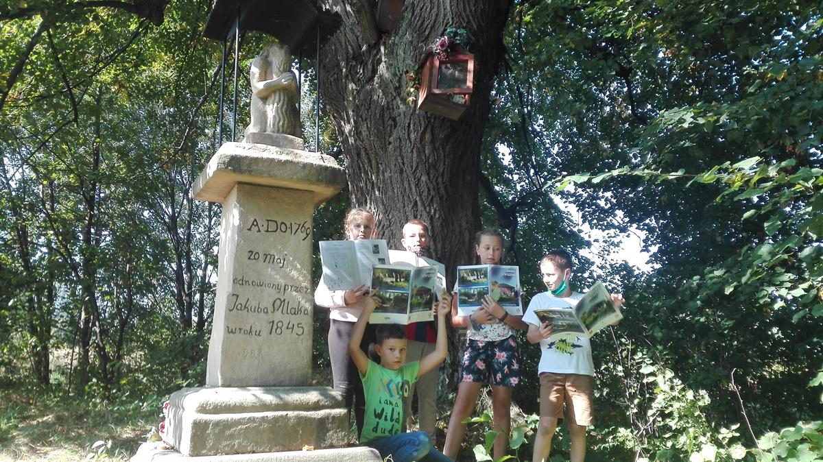 Dzieci ze Stronia stoją obok figury świętego Onufrego trzymając ćwiczenia do lekcji regionalizmu w rękach.