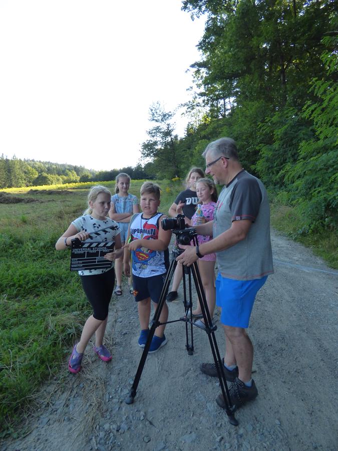 Uczestnicy zajęć filmowych stoją na drodze w plenerze razem z panem instruktorem, który pokazuje im, jak działa aparat fotograficzny.