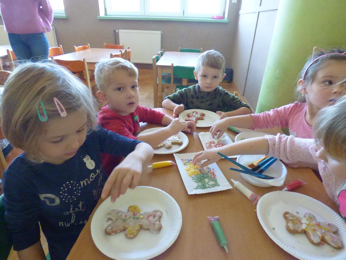Przedszkolaki siedzą przy stoliku, malują lukrem i dekorują pisakami cukrowymi i różnego rodzaju posypkami pierniczki.