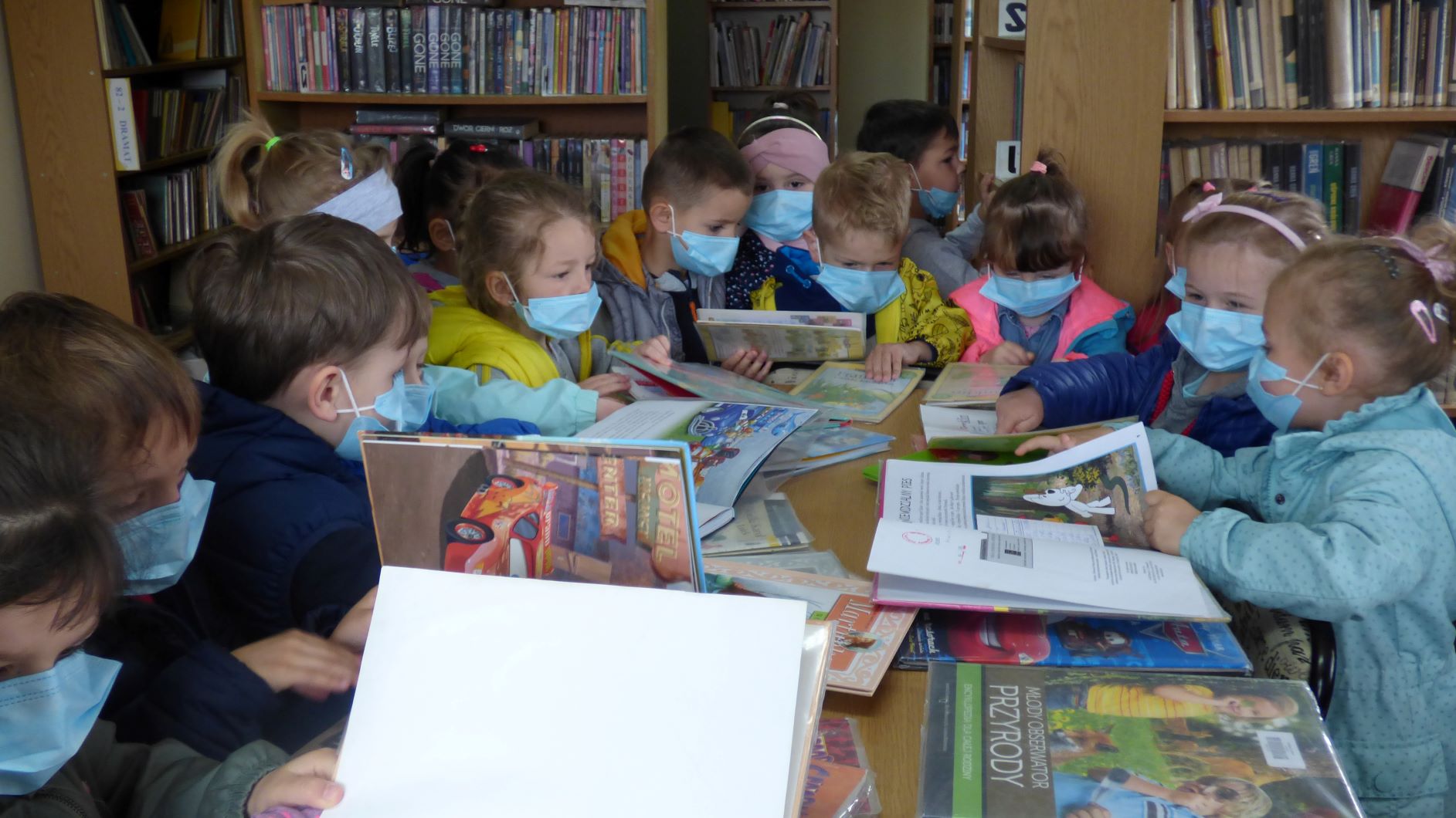 Przedszkolaki stoją wokół stołu w bibliotece i oglądają książeczki dla dzieci.