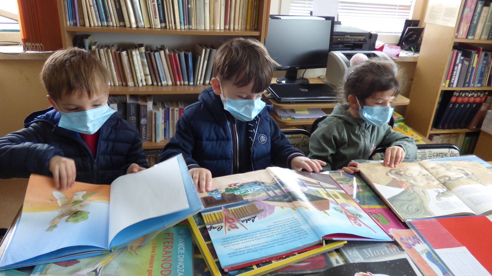 Troje dzieci stoi przy stole i ogląda otwarte książki dla dzieci.