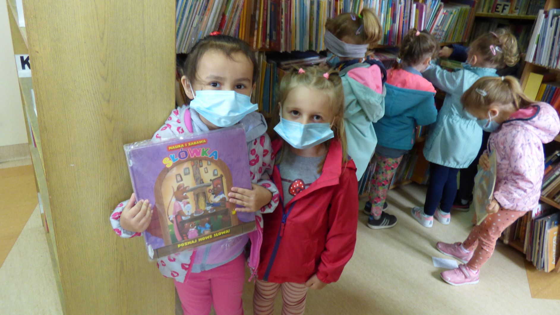 Dwie dziewczynki pozują do zdjęcia patrząc w obiektyw i trzymając w ręce książeczkę. Czworo dzieci z tyłu stoi przy regale z książkami.