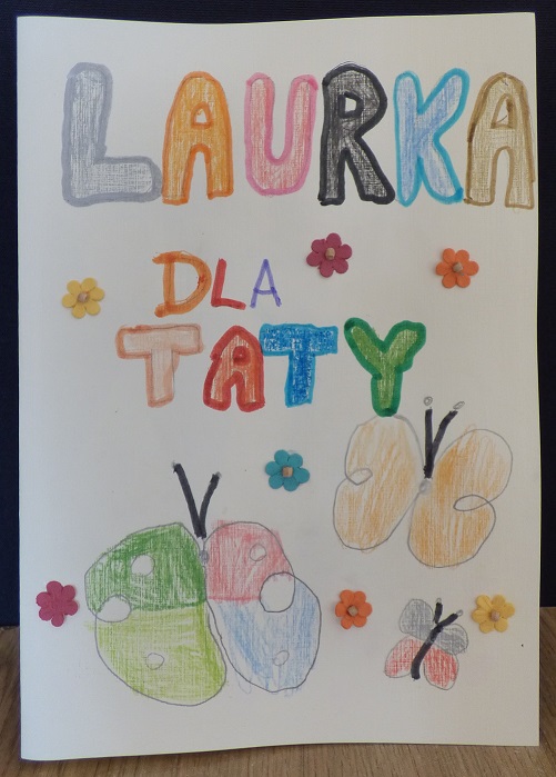 Na środku zdjęcia jest kolorowy napis "Laurka dla Taty" narysowany kredkami, pod napisem narysowane są kolorowe motyle.
