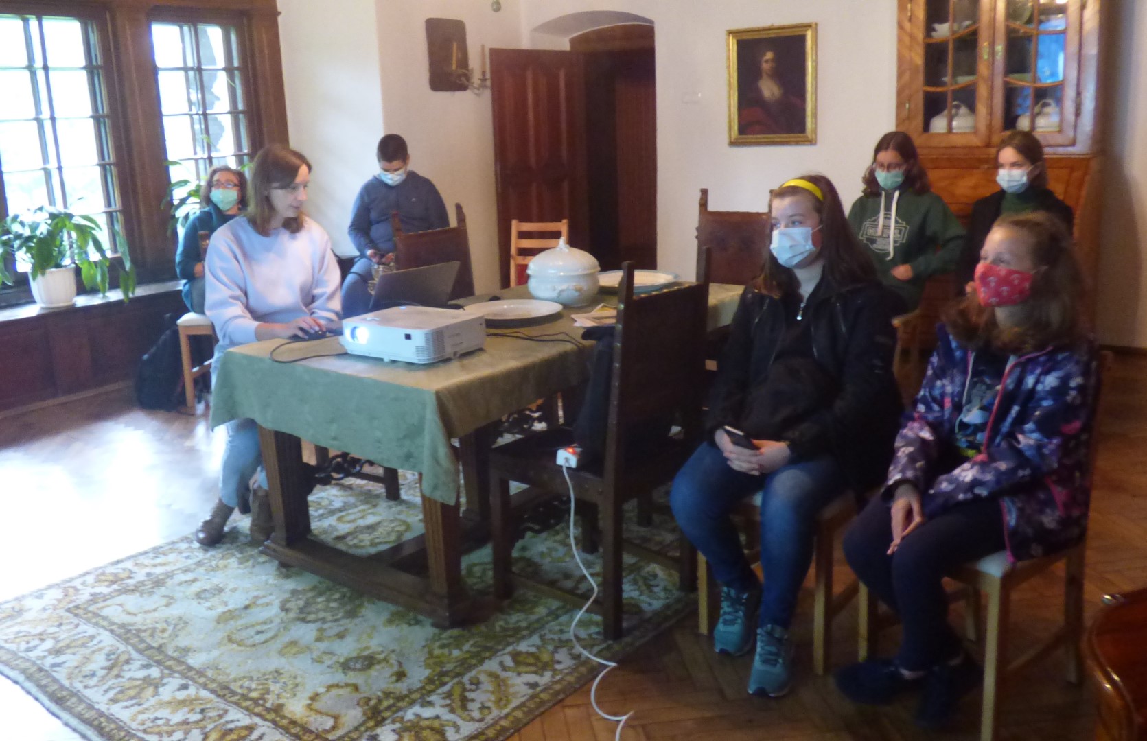 Wokół stołu siedzi pięciu uczestników projektu wraz z instruktorem słuchając historii mieszkańców Dworu w Stryszowie, którą opowiada im pani kierownik tegoż dworu.