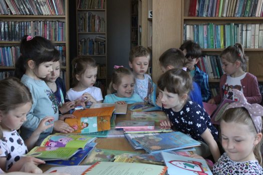 W pomieszczeniu biblioteki publicznej, wokół stołu stoją dzieci z grupy pięciolatków, które oglądają rozłożone na stole książeczki.