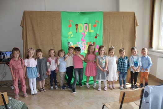 Dzieci z grup przedszkolnych trzylatków, czterolatków i pięciolatków stoi obok siebie na tle zielonej dekoracji i pozuje do zdjęcia po zaśpiewaniu piosenek konkursowych.
