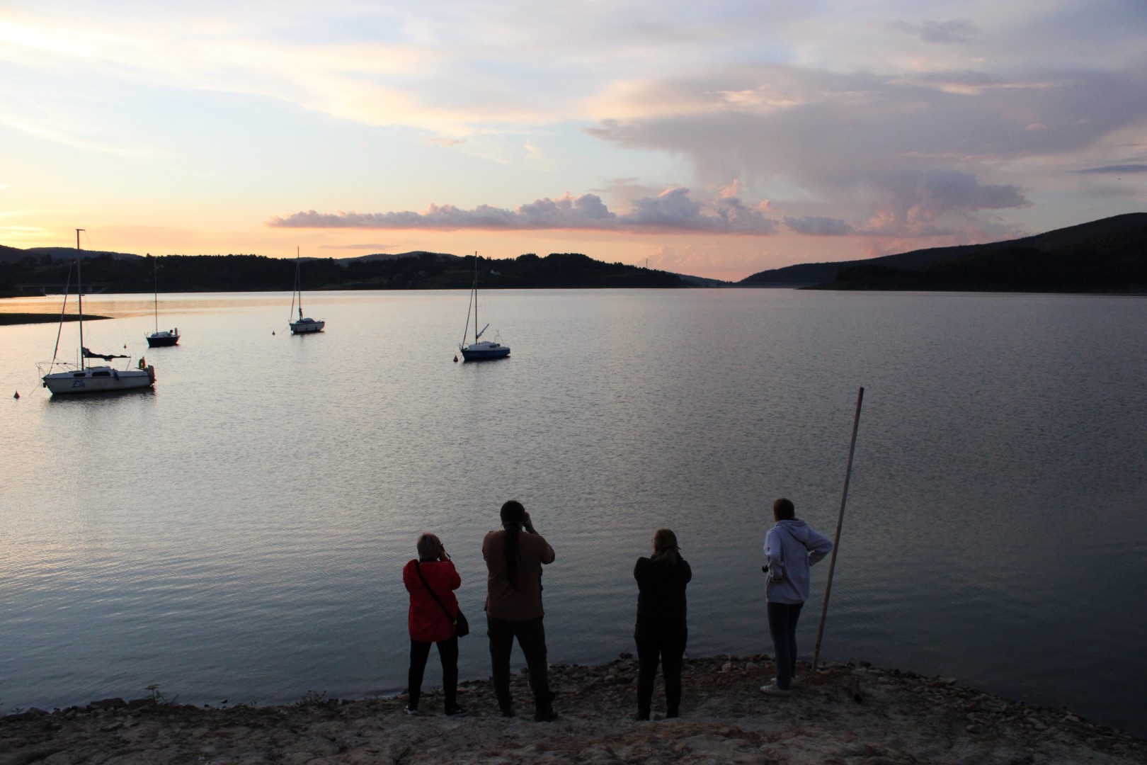 Trzy uczestniczki projektowych warsztatów fotograficznych wraz z instruktorem stoją na brzegu Jeziora Mucharskiego i robią zdjęcia tafli jeziora i żaglówkom.
