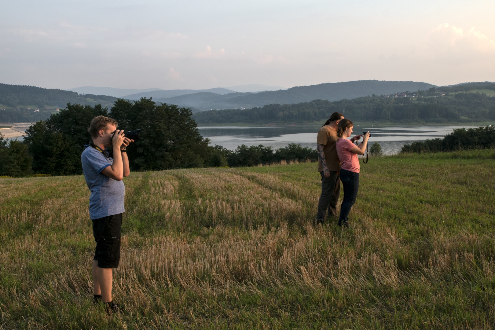 Dwoje uczestników warsztatów fotograficznych wraz z instruktorem fotografem stoją z aparatami w rękach i fotografują przestrzeń przed sobą.