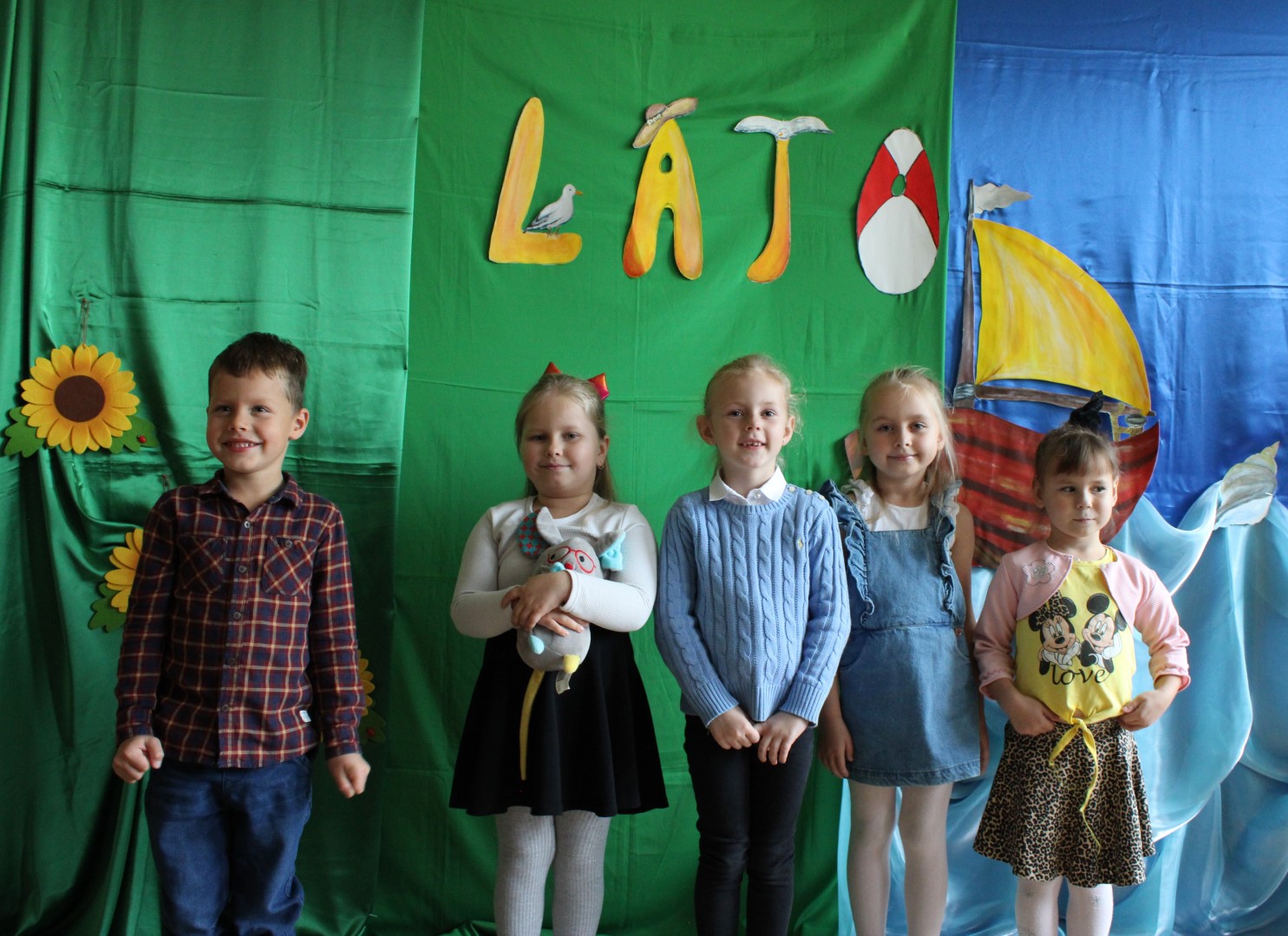 Zdjęcie przedstawia pięcioro dzieci: cztery dziewczynki i jednego chłopca, pozujących do zdjęcia grupowego podczas Konkursu Recytatorskiego „Cztery Pory Roku – LATO” organizowanego przez Gminny Ośrodek Kultury w Stryszowie. Dzieci stoją na tle zielono-niebieskiej dekoracji, nad nimi widnieje żółty napis LATO.
