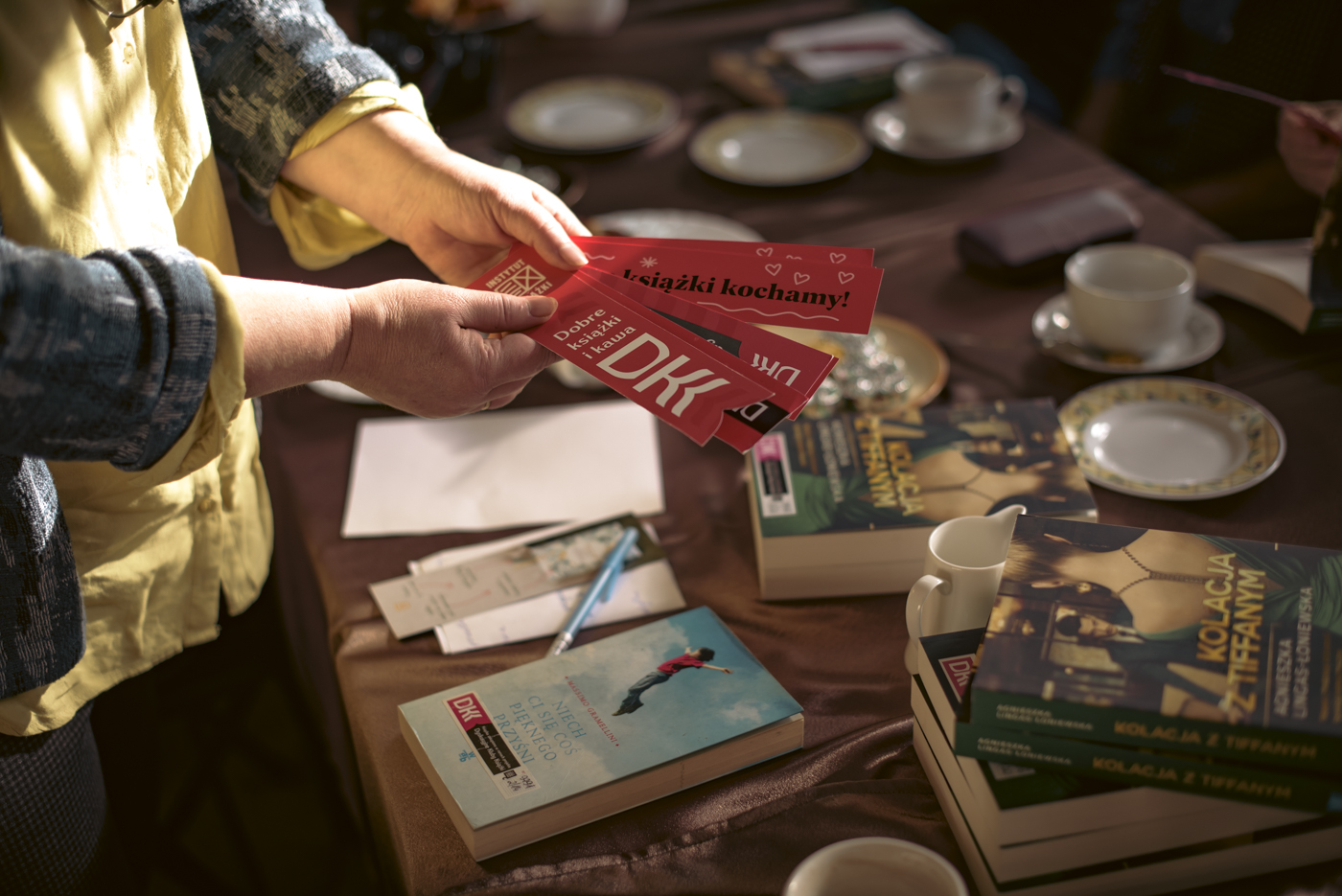 Zdjęcie przedstawia kobietę trzymającą w dłoniach nad stołem zakładki do książek, które są zaprojektowane zgodnie z przedsięwzięciem Dyskusyjnego Klubu Książki, są one czerwone z logo DKK.