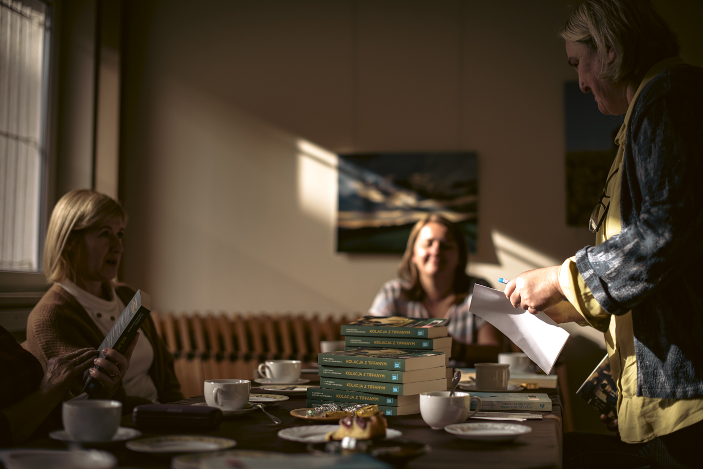 Zdjęcie przedstawia trzy kobiety na świetlicy GOK w Stroniu. Dwie z nich siedzą przy stole, natomiast trzecia stoi i rozdaje nowe książki, które będą omawiane na kolejnym spotkaniu DKK.