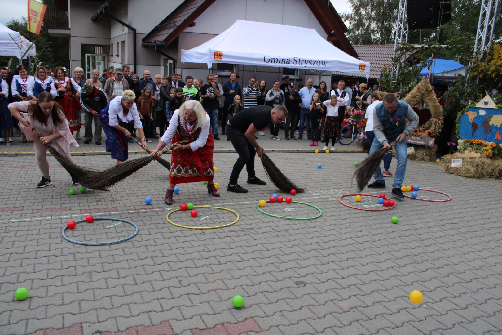 Zdjęcie przedstawia jedną z konkurencji dożynkowych Turnieju Sołectw o Puchar Wójta Gminy Stryszów, podczas której reprezentanci sześciu sołectw zamiatają miotłami piłeczki na wyznaczone miejsce.