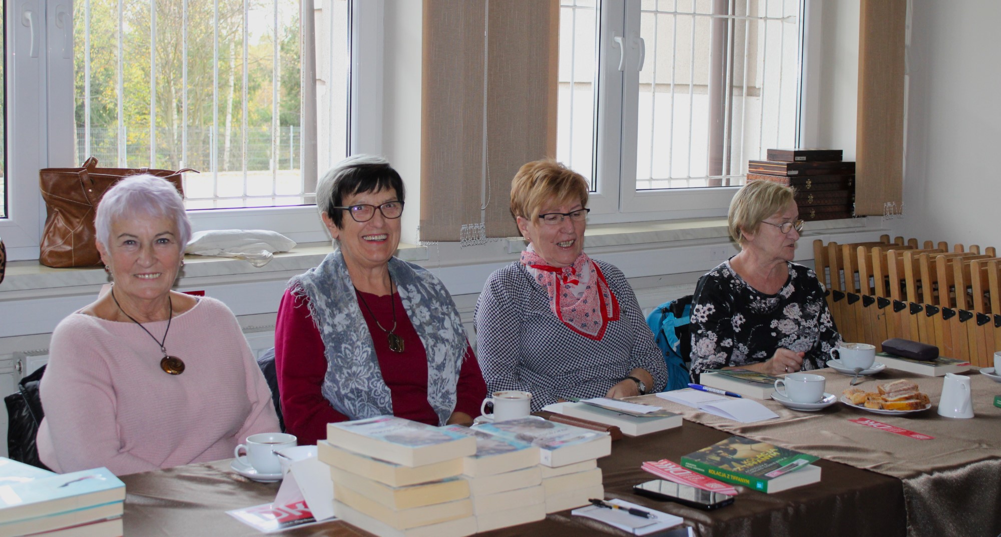 Przy stole siedzą cztery kobiety. Na stole książki i filiżanki z kawą