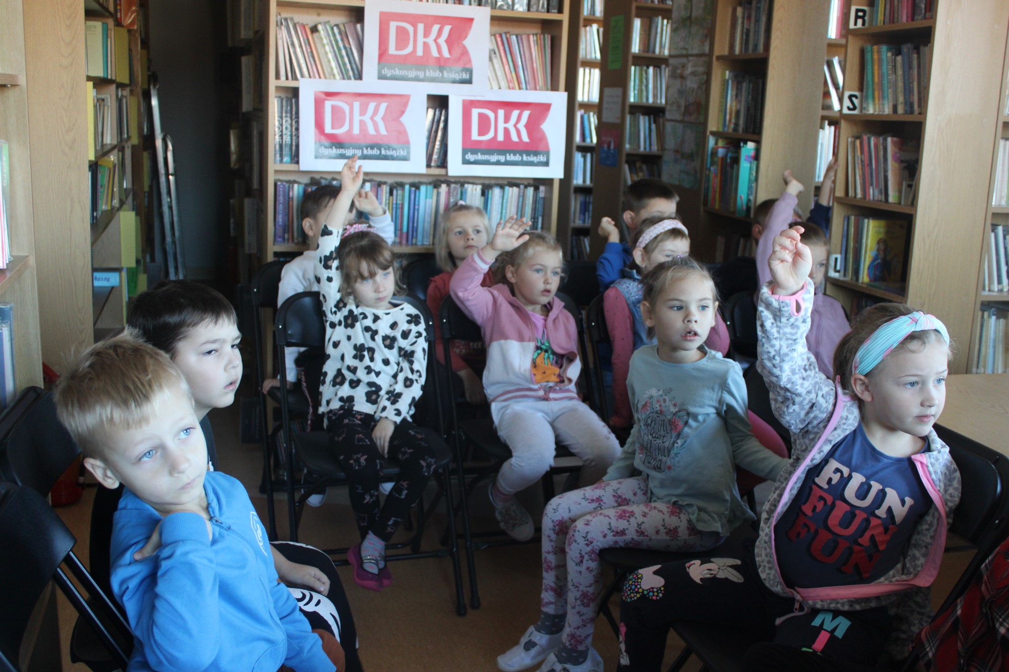 Grupa dwunastu dzieci siedzących na krzesłach, trzyma ręce w górze. Zadają oni pytania Pani Bibliotekarce dotyczące przeczytanej książki „Moje zabawki są moje”, w ramach programu Dyskusyjnego Klubu Książki dla Przedszkolaka prowadzonej w Gminie Stryszów przez Biblioteki Publiczne.