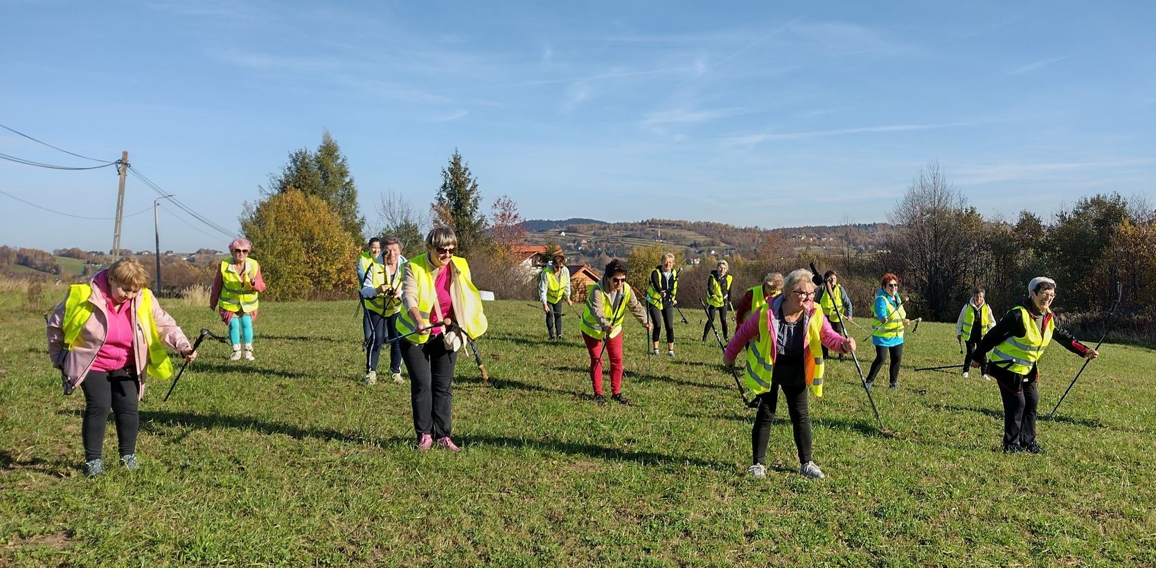 Na zdjęciu znajduje się osiemnaście kobiet, które w rzędach wykonują ćwiczenia rozciągające ich mięśnie, podczas zorganizowanego wspólnego spaceru szlakami Nordic Walking.