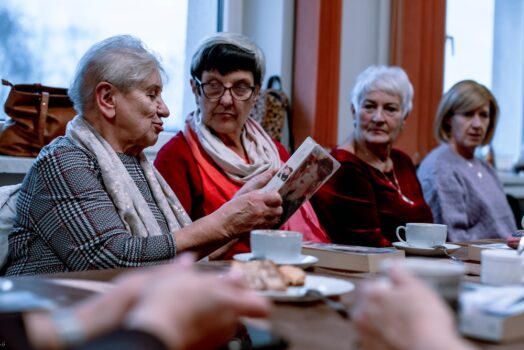 cztery kobiety siedzą przy stole. Jedna z nich trzyma ksiązkę