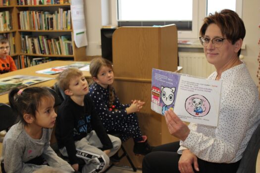 Na zdjęciu znajduje się Pani bibliotekarz z Biblioteki Publicznej w Stroniu, trzyma w ręku książkę, którą pokazuje przedszkolakom siedzącym przed nią na krzesełkach.