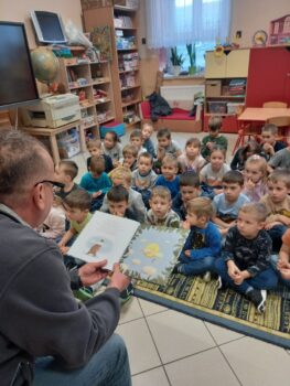 Zdjęcie przedstawia siedzącego na stołku bibliotekarza z Biblioteki Publicznej w Stryszowie, który czyta siedzącym na dywanie dzieciom w przedszkolu Stryszowie książkę pod tytułem „Proszę mnie przytulić” Przemysława Wechterrowicza.