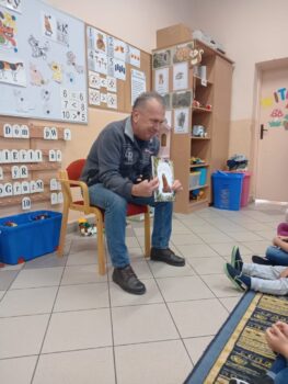 Zdjęcie przedstawia siedzącego na stołku bibliotekarza z Biblioteki Publicznej w Stryszowie, który czyta dzieciom w przedszkolu w Stryszowie książkę pod tytułem „Proszę mnie przytulić” Przemysława Wechterrowicza.